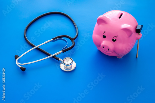 Money for treatment. Medical expenses. Moneybox in shape of pig near stethoscope on blue background copy space