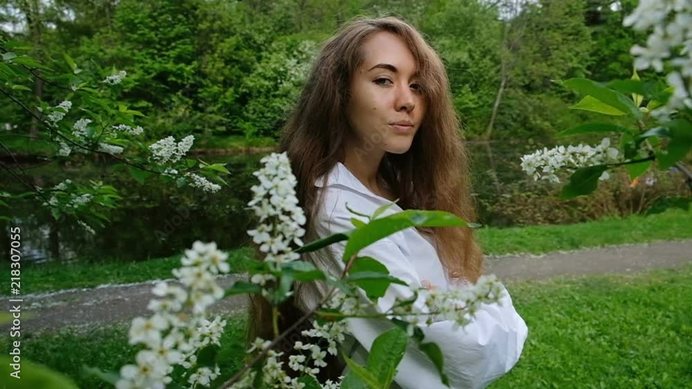 Pretty European girl model posing on camera in blossoming bird cherry. Young woman with long fluffy hair and a branch on her head is photographed in the park, slow motion Pretty European girl model posing on camera in blossoming bird cherry. Young woman with long fluffy hair and a branch on her head is photographed in the park, slow motion