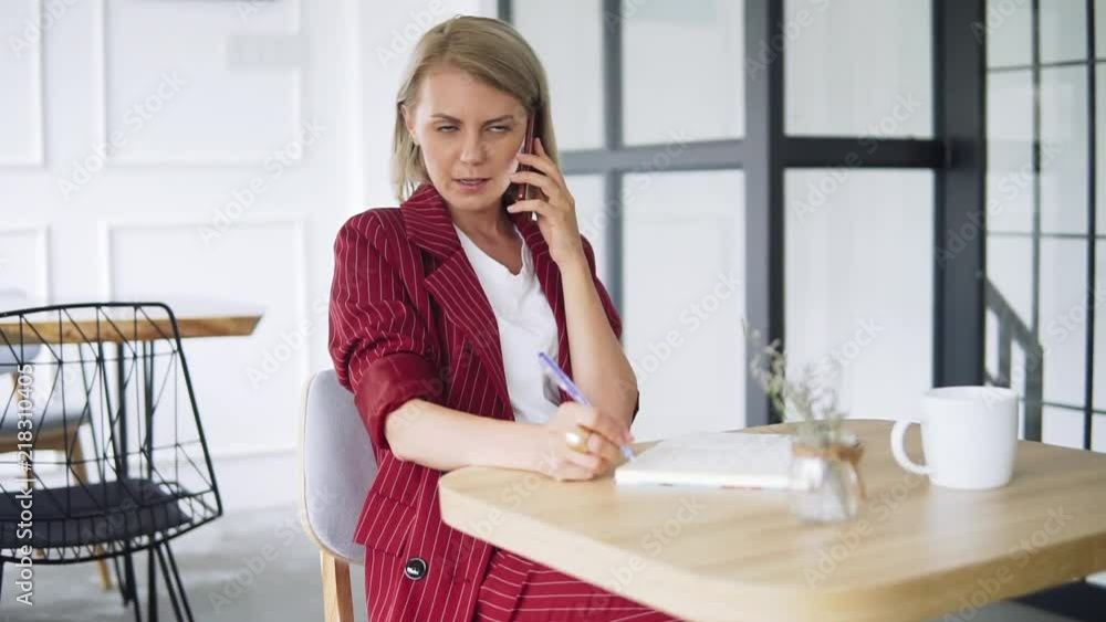 Beautiful and young successful business woman with mobile phone and notepad in a cafe, working as a freelancer. Blonde Female in red formal suit sitting at the table and talking with the friend