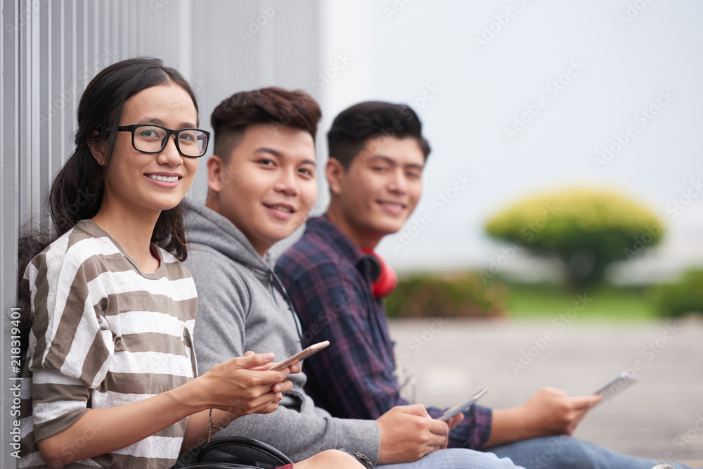 Pretty smiling Vietnamese girl with smartphone resting outdoors with her friends Pretty smiling Vietnamese girl with smartphone resting outdoors with her friends