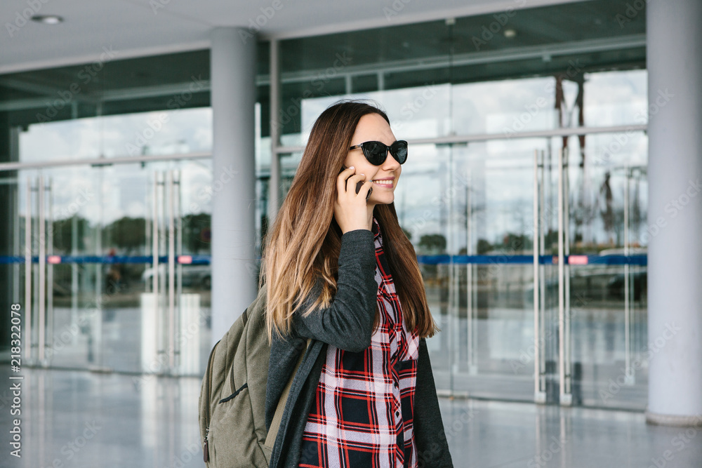 Fototapeta premium Young beautiful tourist girl at the airport or near the shopping center or the station calls a taxi or talking on a cell phone or talking to friends using a mobile phone.