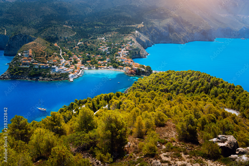 Breathtaking top view to Assos village with local houses. Landmark place of Kefalonia. Lonely white yacht at anchor in calm beautiful lagoon surrounded by pine and cypress trees. Greece