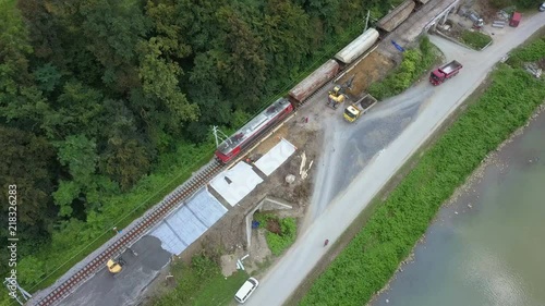 Aerial: Flying over train passing through railway reconstruction site. Filming rails renovation at railway beside beautiful river on sunny day.