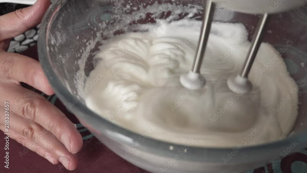 Someone using a blender to finish mounting the egg whites in snow in a transparent salad bowl Someone using a blender to finish mounting the egg whites in snow in a transparent salad bowl