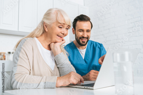 smiling young caregiver and happy senior woman using laptop together