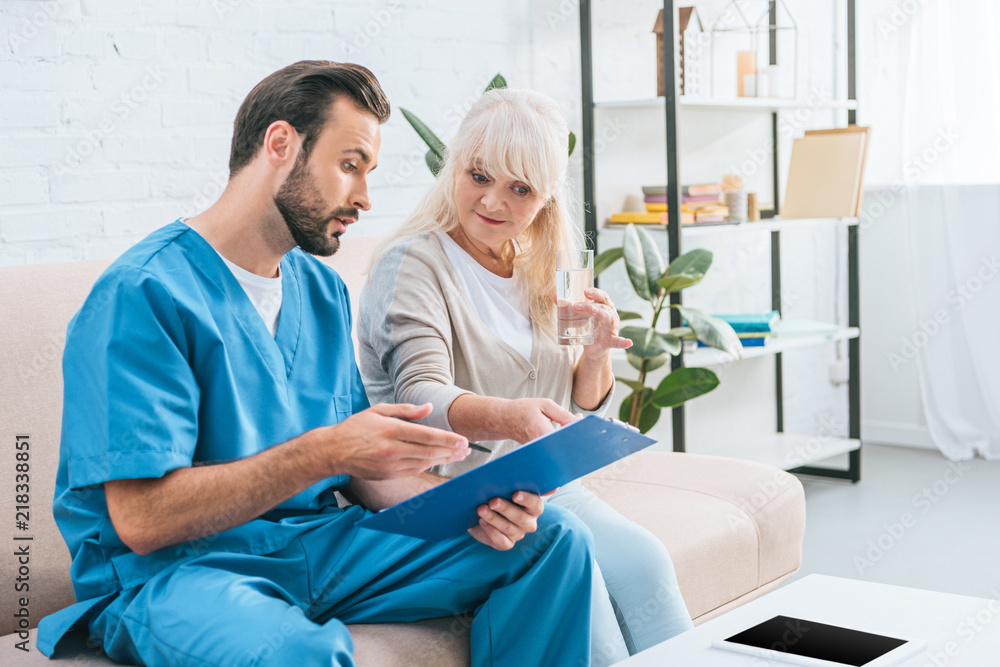 young male nurse showing clipboard to senior woman holding glass of water young male nurse showing clipboard to senior woman holding glass of water