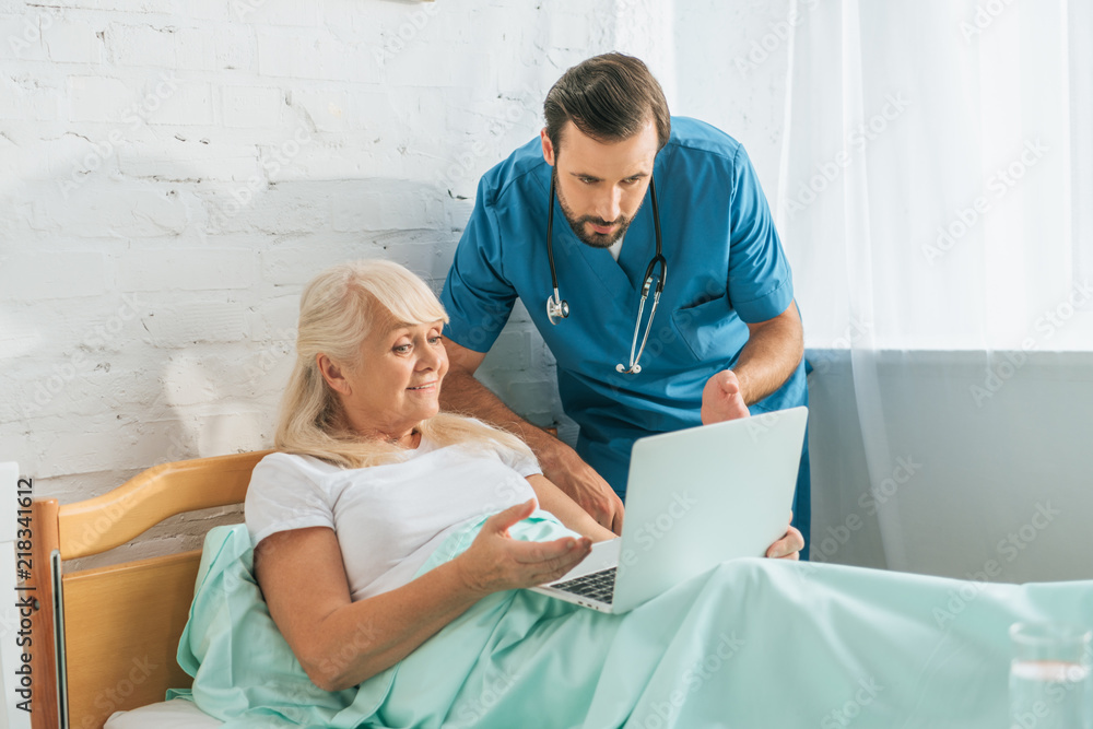 Obraz premium doctor with stethoscope looking at senior woman using laptop in hospital bed