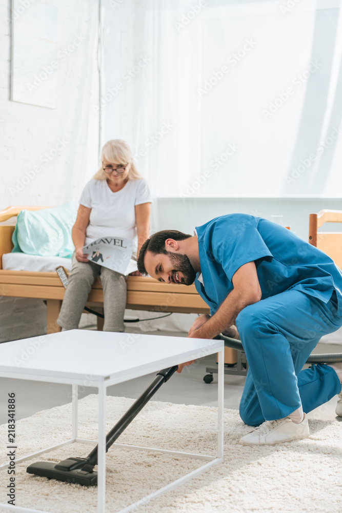young caregiver cleaning carpet with vacuum cleaner while senior woman reading newspaper on bed young caregiver cleaning carpet with vacuum cleaner while senior woman reading newspaper on bed
