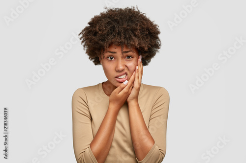Isolated shot of displeased dark skinned young female has Afro haircut, keeps hand on cheek, frowns face from pain, suffers from toothache, dressed in casual sweater, expresses negative emotions photo
