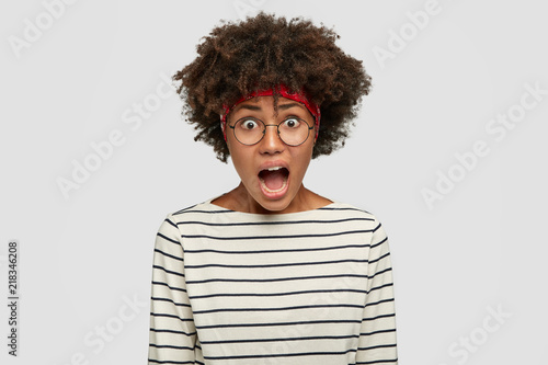 Mad African American female shouts with annoyed expression  has bushy hair  dressed in black and white casual striped sweater  keeps jaw dropped  feels anxious  isolated over white background
