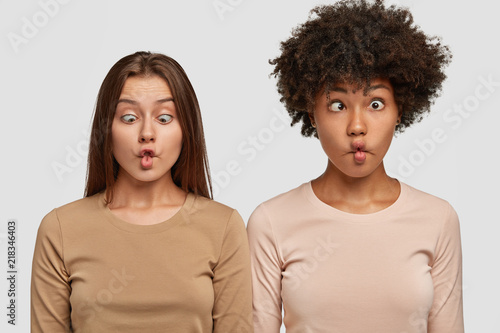 Funny mixed race female friends pout lips, make grimaces and have fun together, being tired after work on project, foolish indoor isolated over white background. People, interracial friendship concept