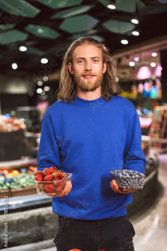 Young smiling guy in dark blue sweater dreamily looking in camera while holding blueberries and strawberries in hands in modern supermarket