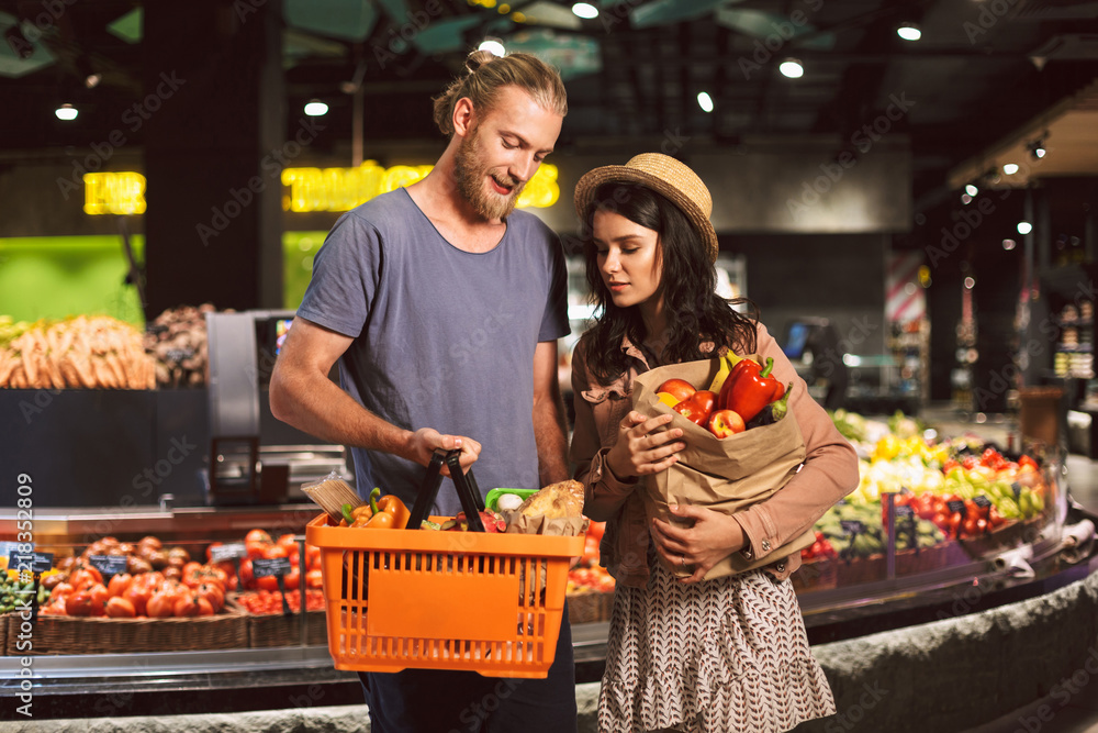 Young guy and girl dreamily looking on basket and paper grocery bag full of products in supermarket