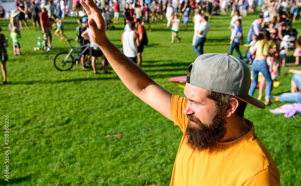 Naklejka premium Man bearded hipster in front of crowd people green field background. Urban event celebration. Man waving hand sunny day outdoors. Hipster in cap visiting social event picnic fest or festival
