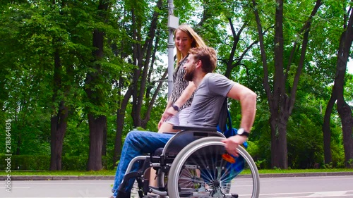 Dolly shot. Happy young disable man in wheelchair walking in the park with his wife. He holding her hand. They are talking and smiling. Side view.