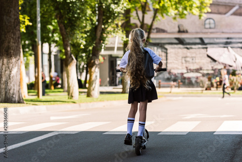 rear view of cute little kid with backpack riding scooter on street