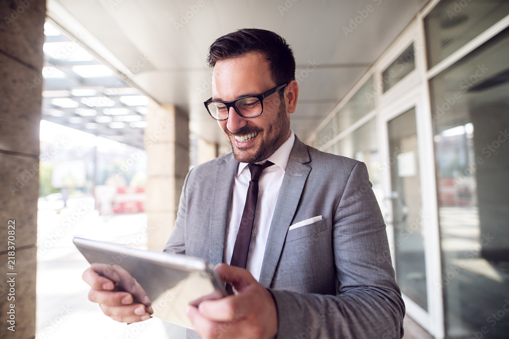 Young modern businessman standing outside looking at his digital tablet with satisfaction on his face.Good day at job. Young modern businessman standing outside looking at his digital tablet with satisfaction on his face.Good day at job.