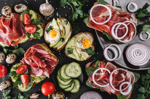 Various of sandwiches and bruschetta with prosciutto, fried quail egg, avocado, cucumber, tomatoes, spices and greens on black stone background. Clean eating, healthy breakfast. Top view, flat lay