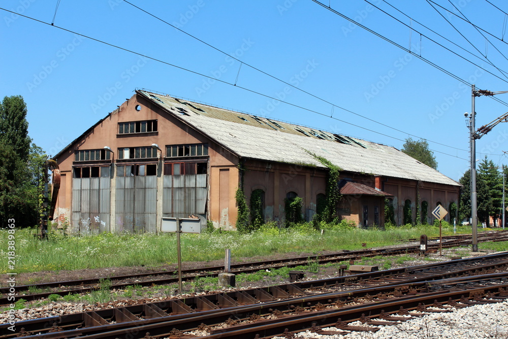 Dilapidated abandoned railway station building with missing roof tiles and windows surrounded with tall uncut grass and other vegetation next to rusted railway tracks with clear blue sky in background Dilapidated abandoned railway station building with missing roof tiles and windows surrounded with tall uncut grass and other vegetation next to rusted railway tracks with clear blue sky in background
