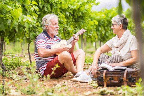 couple of alternative aged older traveler stay sitting down in a vineyard with the luggage and playing an ukulele acoustic guitar and reading a paper book. easy lifestyle in outdoor places