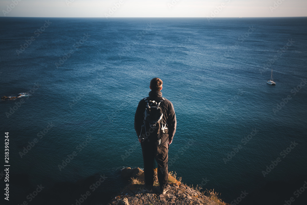 A rear view of a male backpacker or hiker standing on a high clifftop and overlooking a vast ocean at Kynance Cove in Cornwall, UK