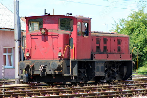 Old used dark red electric locomotive parked on railway tracks waiting for departure from local railway station with trees and blue sky in background