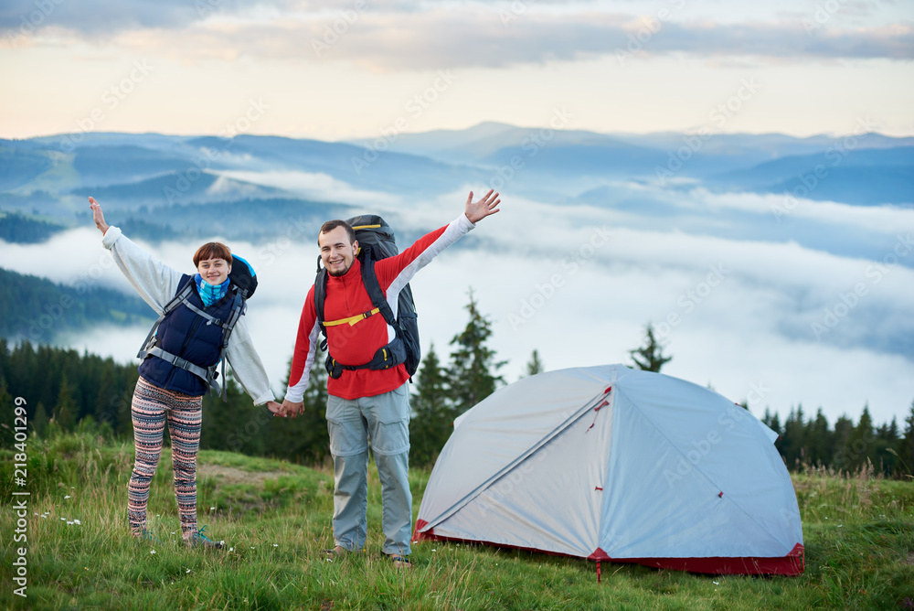 Fototapeta premium Happy couple with backpacks near the tent holding hands against the backdrop of the beautiful scenery of the mighty mountains of the Carpathians on which lies a haze and the sky is beautiful clouds.