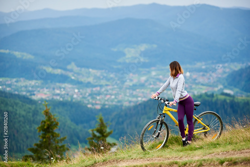 Athlete young woman cyclist with her yellow bicycle on a rural trail enjoying evening view of mountains, forests and small city on the blurred background. Outdoor sport activity. Copy space