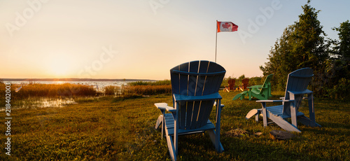 Muskoka chairs on a lawn beside a lake with a Canadian flag in the background at sunset. photo