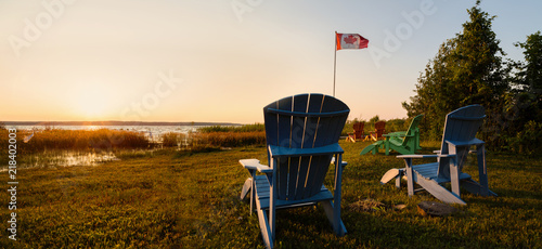 Muskoka chairs on a lawn beside a lake with a Canadian flag in the background at sunset.