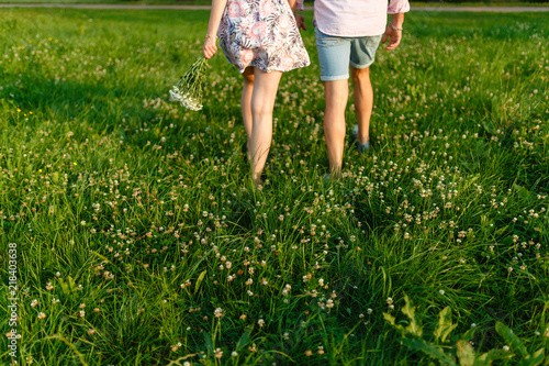 Rear view of a romantic man and woman stand on walk on field grass. Concept of lovely family holding hands. Young couple running through a green meadow. A girl in a summer dress and a guy in shorts.