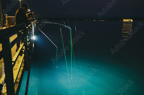 Squid fishermen extend their poles into the lit water below a pier