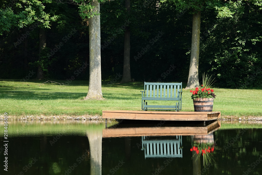 Obraz premium Empty Bench and Geraniums in Barrel Beside Pond for Quiet Reflection