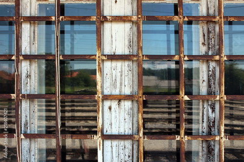 Rusted metal bars in shape of net mounted on old dilapidated white wooden window frame with windows reflecting houses  trees and clear blue sky in background