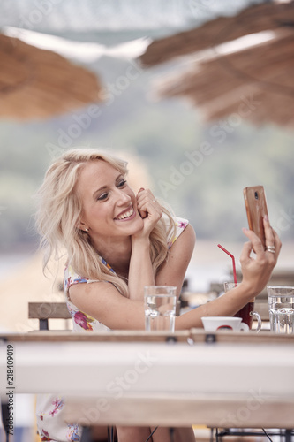 one young beautiful woman smiling, 25 years old, smartphone selfie self-portrait, sitting in beach cafe, summer sunshades table chair.