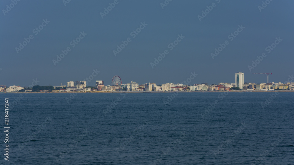 View from the sea on the italian city on the coastline with skyscrapers and ferris wheel. 