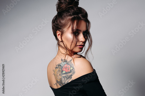 Pretty girl with gathered dark brown hair wearing black casual blouse with opened shoulders showing colorful tattoo. Having big circle earrarings, small nose, big eyes, plump lips. Posing at studio.