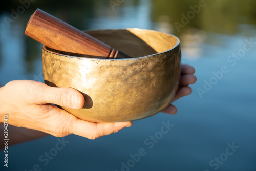 The old singing bowl in the male hands against the background of blue water in the rays of the setting sun.