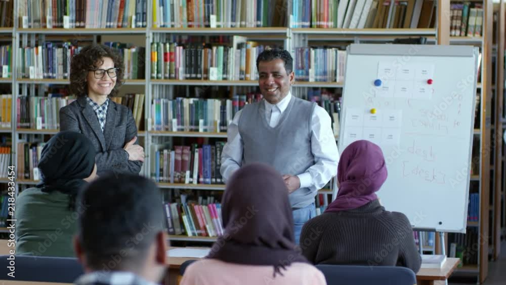 Tilt down of middle eastern man and Caucasian female teacher standing by flipchart in classroom, smiling and talking with group of migrant students Tilt down of middle eastern man and Caucasian female teacher standing by flipchart in classroom, smiling and talking with group of migrant students