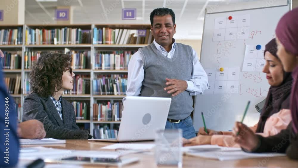 Mid-aged arab man standing at flipchart and explaining English grammar to group of migrant students Mid-aged arab man standing at flipchart and explaining English grammar to group of migrant students