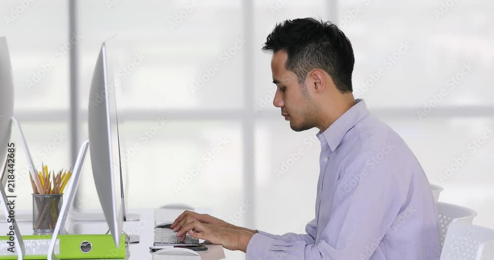 Young and handsome Asian businessman typing on computer keyboard alone in front of new computers in modern office Young and handsome Asian businessman typing on computer keyboard alone in front of new computers in modern office
