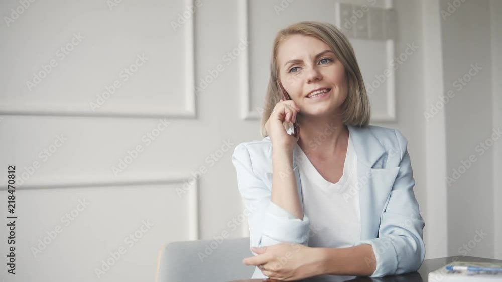 Beautiful and young successful business woman with mobile phone and notepad in a cafe, working as a freelancer. Blonde Female in blue formal suit sitting at the table and talking with the friend Beautiful and young successful business woman with mobile phone and notepad in a cafe, working as a freelancer. Blonde Female in blue formal suit sitting at the table and talking with the friend