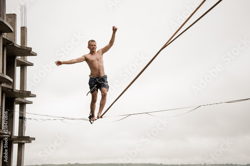 Strong brave man balancing on a slackline high against empty building and sky