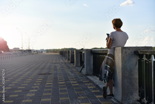woman alone on city embankment hold telephone and looking on sunset