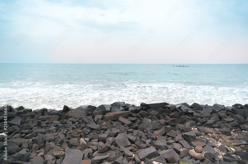 Rocky beach with black brown stones and waves dashing into them. Shot at sunset during a cloudy day with the red sky of sunset. This is promenade beach in pondicherry a famous tourist spot