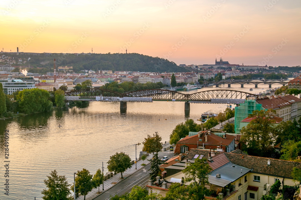 View from Vysehrad of Prague castle, sunset sky with pink and yellow colors, railway bridge, stone bridge, river Vltava, boats, riverbank, streets, cars, houses, red roofs, green trees, hill of Petrin
