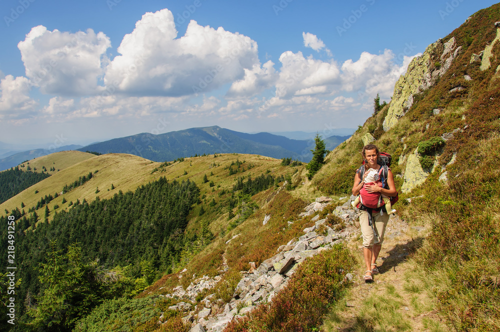 A girl with baby in a sling goes at the mountain range. Ukraine. Carpathian mountains