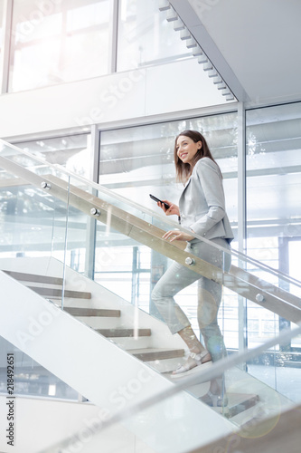 Young smiling businesss woman walking up  the stairs  of a public building  and using a smartphone. Business, technology and communication concept