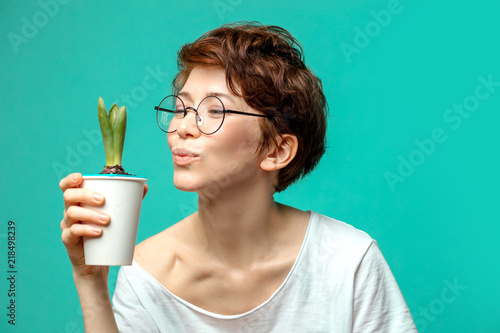 Adorable, a little bit shy hipster woman wearing spactacles and white t-shirt posing isolated with positive expression, holding in hands flower in pot photo