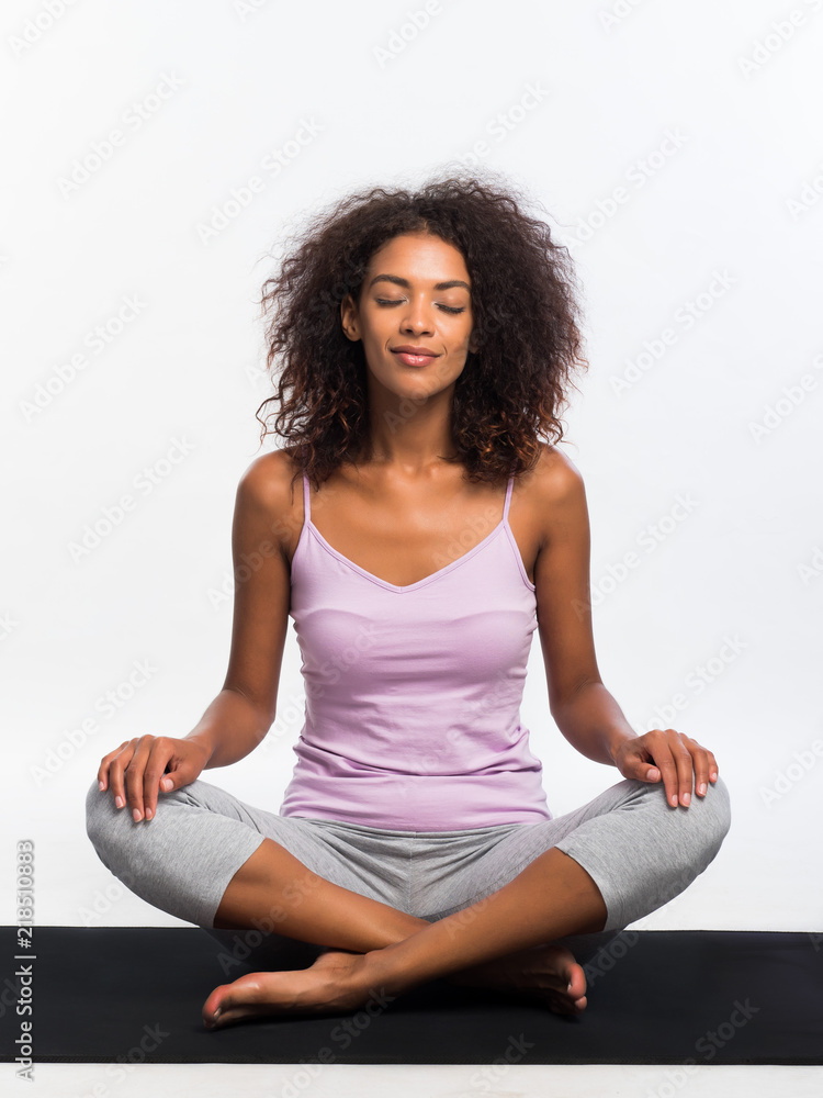 Happy african american woman in comfortable sports wear meditating on mat over white wall background. Black girl concentrated on yoga practice. Happy african american woman in comfortable sports wear meditating on mat over white wall background. Black girl concentrated on yoga practice.